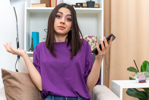 Jong meisje in vrijetijdskleding die een smartphone vasthoudt en er verward uitziet met een spreidende arm naar de zijkant zittend op een bank in een lichte woonkamer living