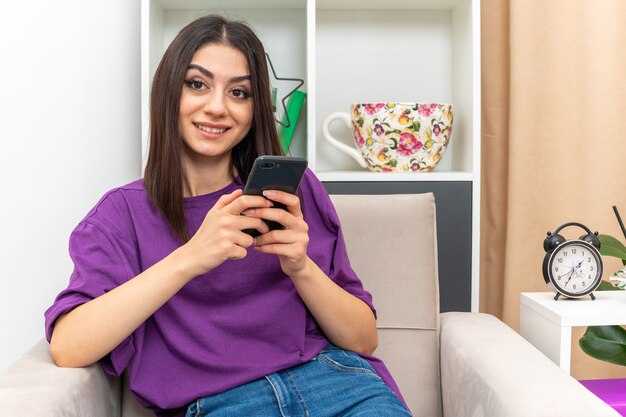 Jong meisje in vrijetijdskleding met smartphone die met een glimlach op het gezicht kijkt, gelukkig en positief zittend op een stoel in een lichte woonkamer living