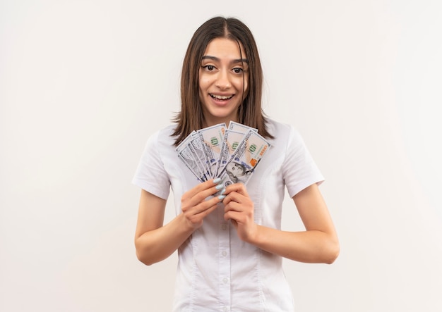 Jong meisje in wit overhemd dat contant geld toont dat vrolijk met gelukkig gezicht glimlacht dat zich over witte muur bevindt