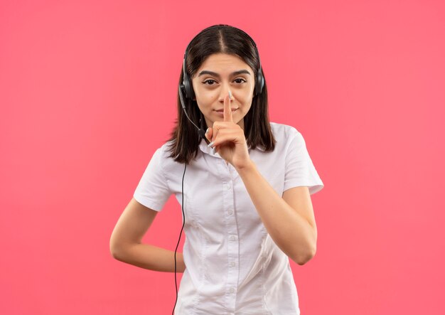 Jong meisje in wit overhemd en hoofdtelefoons, die stilte gebaar met vinger op lippen maken die zich over roze muur bevinden