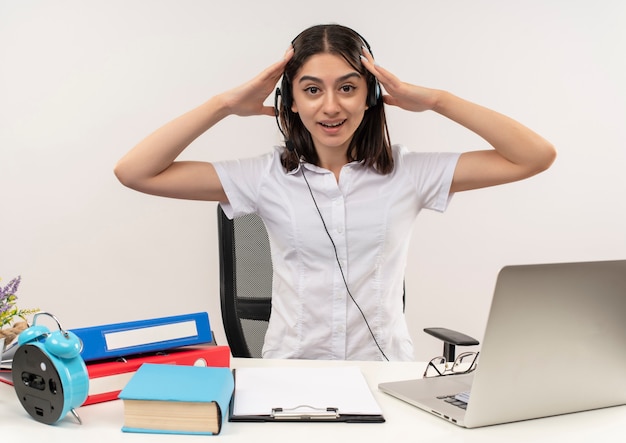 Jong meisje in wit overhemd en koptelefoon, houdt haar hoofd met handen op zoek verward zittend aan tafel met mappen en laptop over witte muur