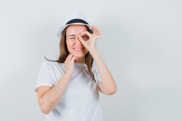 Jong meisje in wit t-shirt, hoed doet ok teken op oog en kijkt vrolijk, vooraanzicht.