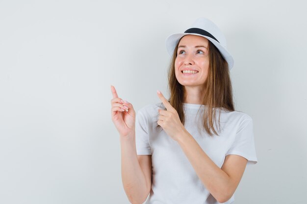 Jong meisje in wit t-shirt, hoed wijst naar de linkerbovenhoek en kijkt vrolijk, vooraanzicht.