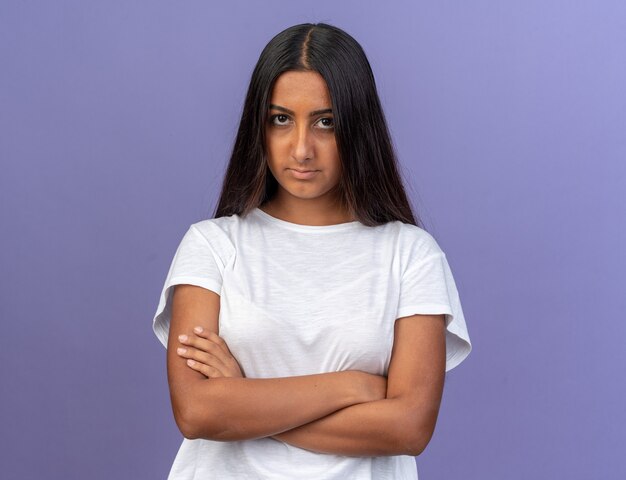 Jong meisje in wit t-shirt kijkend naar camera met ernstig fronsend gezicht met gekruiste armen over blauwe achtergrond