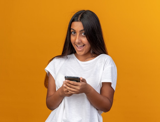 Jong meisje in wit t-shirt met smartphone die naar de camera kijkt met een blij gezicht dat vrolijk lacht smiling