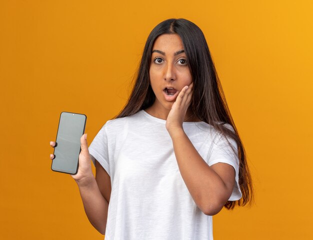 Jong meisje in wit t-shirt met smartphone die verbaasd en verbaasd naar de camera kijkt terwijl ze over oranje achtergrond staat