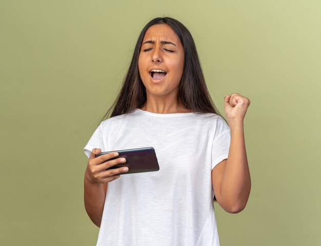 Jong meisje in wit t-shirt met smartphone die vuist opheft, blij en opgewonden over groene achtergrond