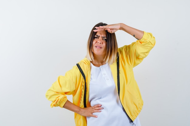 Jong meisje kijkt ver weg met hand boven het hoofd, houdt hand op taille in wit t-shirt, geel jasje en kijkt gefocust. vooraanzicht.