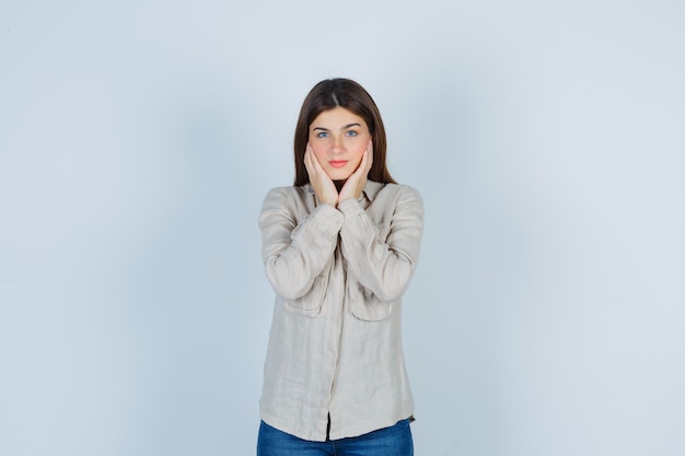 Jong meisje kussen gezicht op handen in beige shirt, jeans en ziet er mooi uit, vooraanzicht.