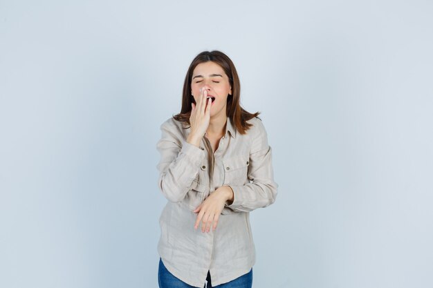 Jong meisje met de hand op de mond terwijl ze in een beige shirt, spijkerbroek gaapt en er slaperig uitziet. vooraanzicht.