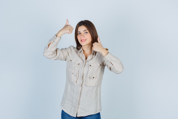 Jong meisje met dubbele duimen omhoog in beige shirt, jeans en vrolijk, vooraanzicht.