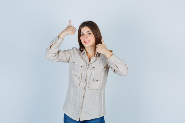 Jong meisje met dubbele duimen omhoog in beige shirt, jeans en vrolijk, vooraanzicht.