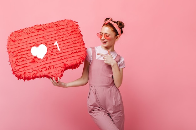 Gratis foto jong meisje met een roze bril met een glimlach kijkt in de camera en wijst naar een leuk teken op een geïsoleerde achtergrond