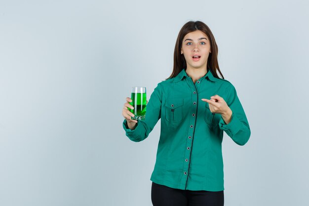 Jong meisje met glas groene vloeistof, erop wijzend met wijsvinger in groene blouse, zwarte broek en verbaasd kijken. vooraanzicht.