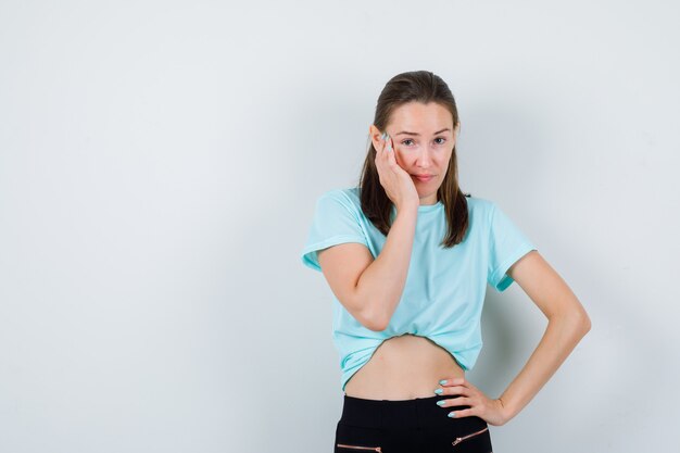 Jong meisje met hand op gezicht in turquoise t-shirt, broek en weemoedig, vooraanzicht.