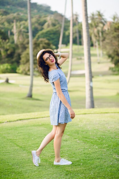 Jong meisje met hemelsblauwe jurk staat in een tropisch park. Het meisje heeft een strohoed en een zwarte bril op.