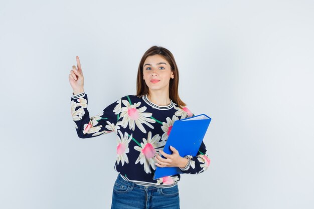 Jong meisje met map, omhoog wijzend in bloemenblouse, jeans en nieuwsgierig kijkend, vooraanzicht.