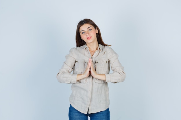 Jong meisje met namaste gebaar in beige shirt, spijkerbroek en schattig, vooraanzicht.