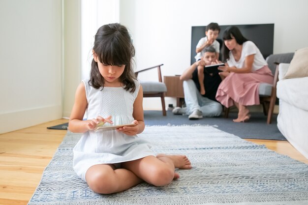 Jong meisje met smartphone zittend op de vloer in de woonkamer, spel terwijl haar ouders en broer met behulp van digitaal apparaat