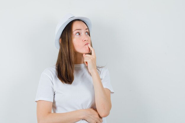 Jong meisje met vinger in de buurt van mond in wit t-shirt, hoed en op zoek naar besluiteloos, vooraanzicht.