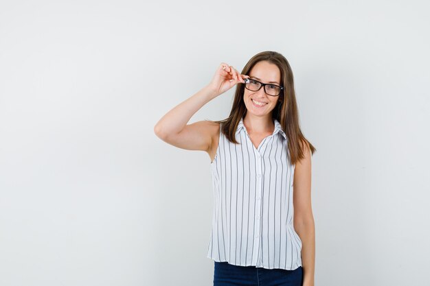 Jong meisje met vingers op bril in t-shirt, spijkerbroek en op zoek joviaal, vooraanzicht.