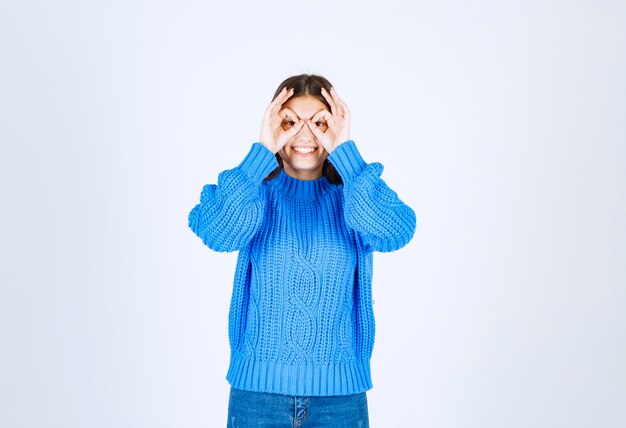 jong meisje model in blauwe trui kijken door vingers op wit-grijs.