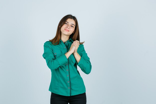 Jong meisje omklemt overhandigt borst in groene blouse, zwarte broek en kijkt gelukkig, vooraanzicht.