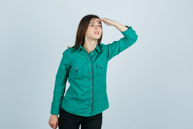 Jong meisje op zoek ver weg met hand boven het hoofd in groene blouse, zwarte broek en gefocust op zoek. vooraanzicht.