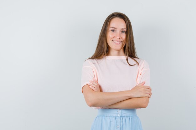 Jong meisje permanent met gekruiste armen in t-shirt, rok en op zoek vrolijk, vooraanzicht.