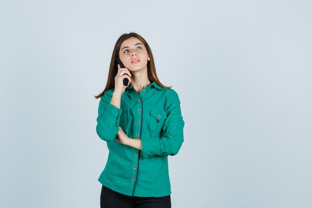 Jong meisje praat met telefoon, kijkend naar boven in groene blouse, zwarte broek en op zoek gericht, vooraanzicht.