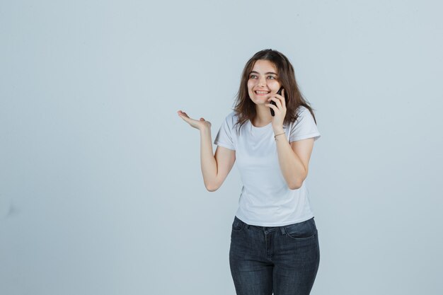 Jong meisje praten op een mobiele telefoon terwijl ze doen alsof ze iets in een t-shirt, spijkerbroek vasthoudt en er vrolijk uitziet. vooraanzicht.