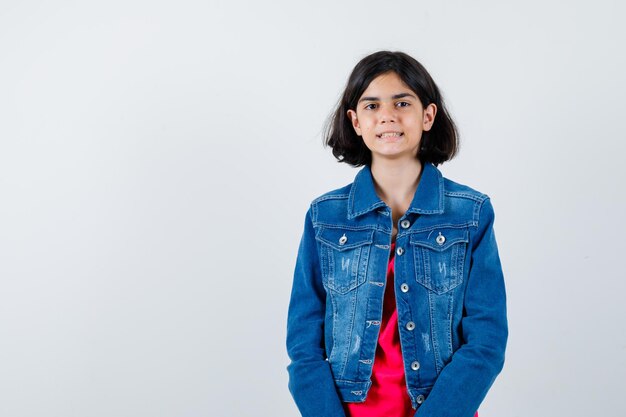 Jong meisje staat rechtop en poseert voor de camera in een rood t-shirt en spijkerjack en ziet er optimistisch uit. vooraanzicht.