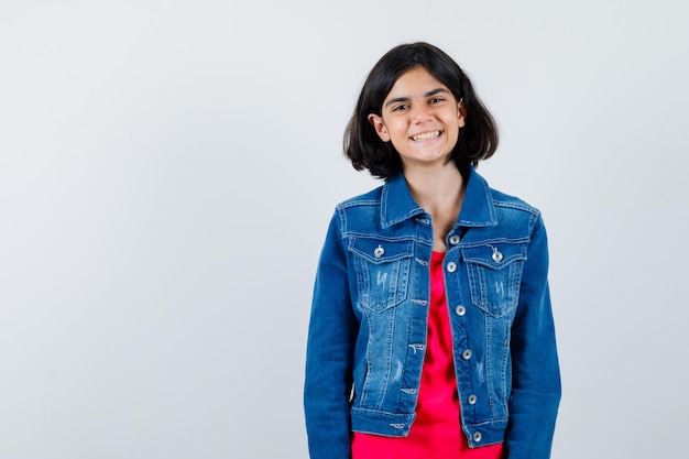 Jong meisje staat rechtop en poseert voor de camera in een rood t-shirt en spijkerjasje en ziet er gelukkig uit. vooraanzicht.