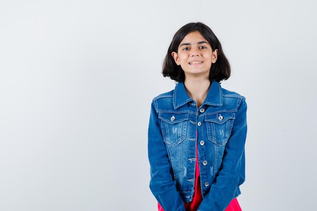 Jong meisje staat rechtop en poseert voor de camera in een rood t-shirt en spijkerjasje en ziet er gelukkig uit. vooraanzicht.