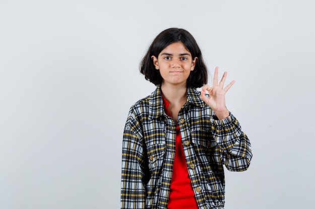 Jong meisje toont ok teken in geruit overhemd en rood t-shirt en ziet er schattig uit. vooraanzicht.