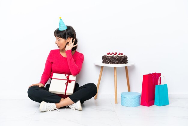 Jong meisje viert zijn verjaardag zittend op de vloer geïsoleerd op een witte achtergrond luisteren naar iets door hand op het oor te leggen