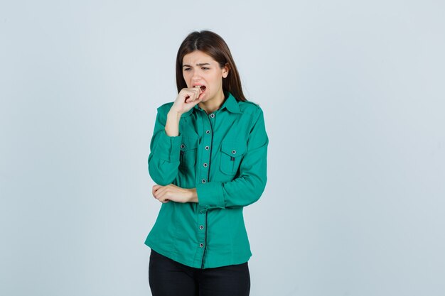 Jong meisje vingers emotioneel bijten in groene blouse, zwarte broek en op zoek bezorgd. vooraanzicht.