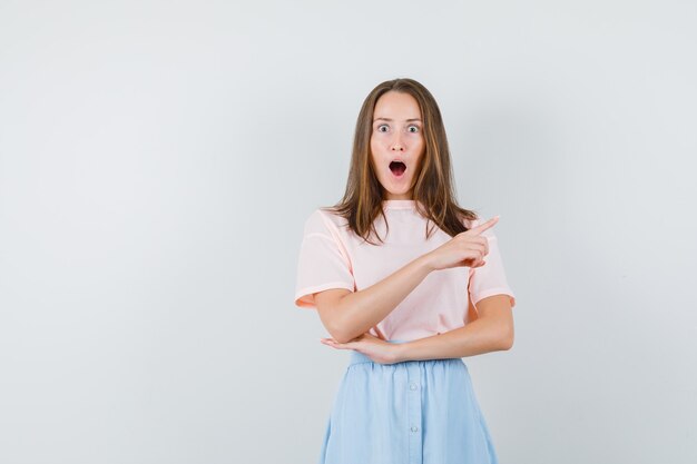 Jong meisje wijst naar kant in t-shirt, rok en kijkt verbaasd, vooraanzicht.