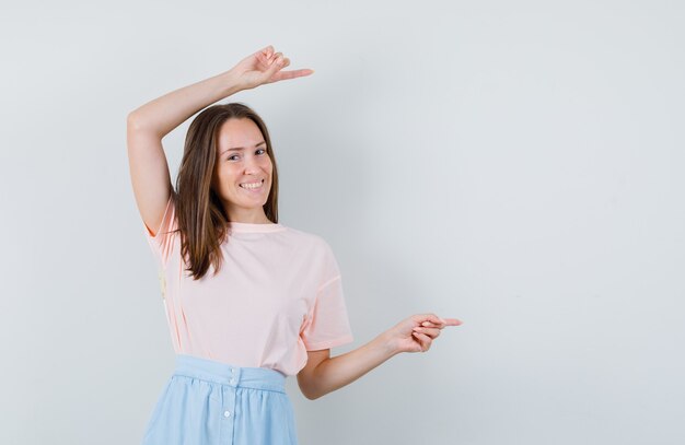 Jong meisje wijst naar kant in t-shirt, rok en kijkt vrolijk. vooraanzicht.