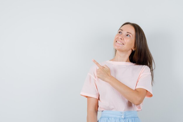 Jong meisje wijst naar kant in t-shirt, rok en kijkt vrolijk, vooraanzicht.