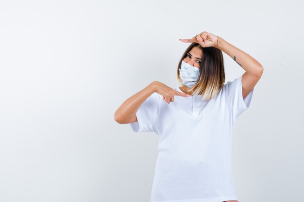 Jong meisje wijst naar rechts en links met wijsvingers in wit t-shirt en masker en kijkt zelfverzekerd, vooraanzicht.
