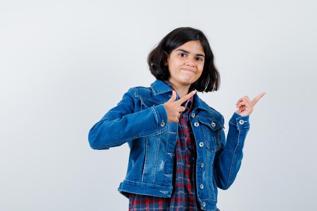 Jong meisje wijst naar rechts met wijsvingers in geruit overhemd en spijkerjasje en ziet er schattig uit