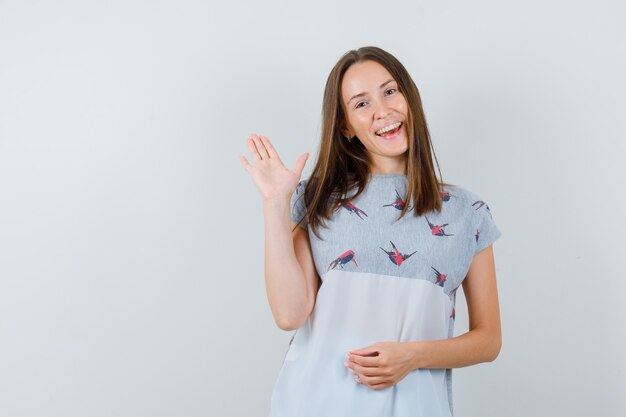 Jong meisje zwaaiende hand voor groet in t-shirt en op zoek vrolijk. vooraanzicht.