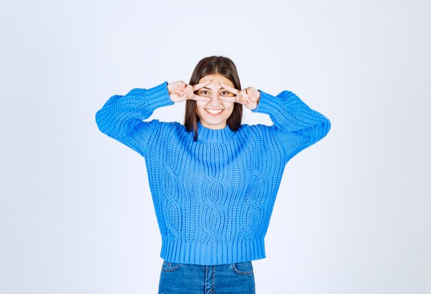 jong meisjesmodel in blauwe sweater die twee vingers dichtbij ogen toont.