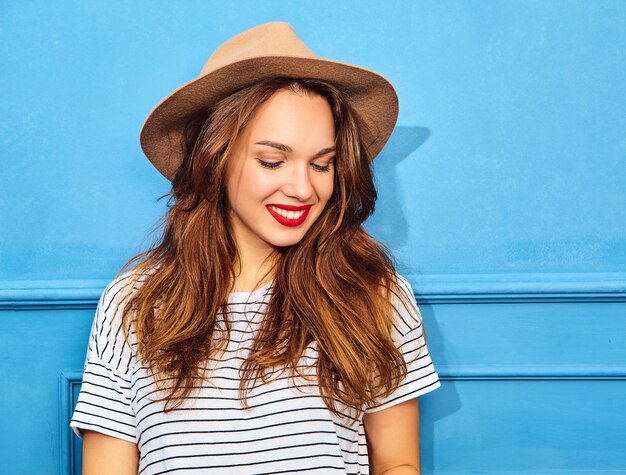 Jong modieus vrouwenmodel in toevallige de zomerkleren en bruine hoed met rode lippen, die dichtbij blauwe muur stellen