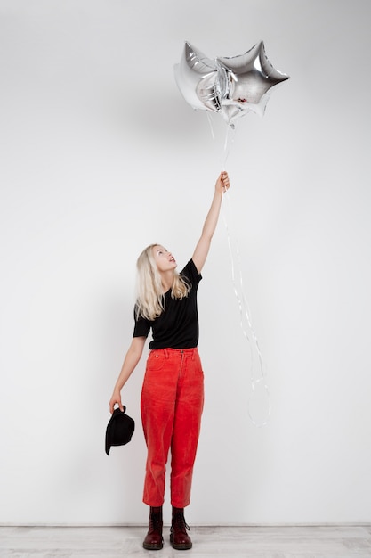 Jong mooi blondemeisje die zilveren baloons over witte muur houden.