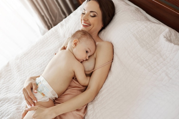 Jong mooi gelukkig mamma die de koesterend haar baby de borst geven die thuis op bed liggen.