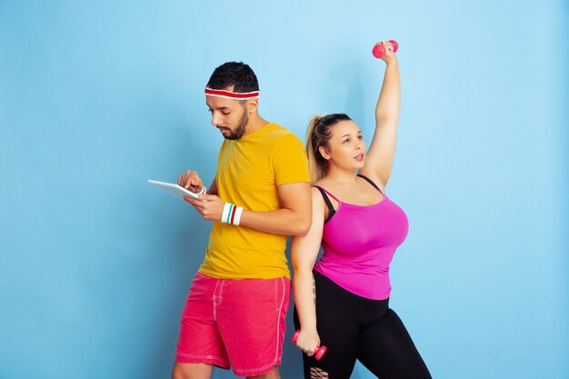 Jong mooi Kaukasisch paar in lichte kleren opleiding op blauwe achtergrond Concept van sport, menselijke emoties, meningsuiting, gezonde levensstijl, relatie, familie. Ze traint, hij gebruikt een tablet.