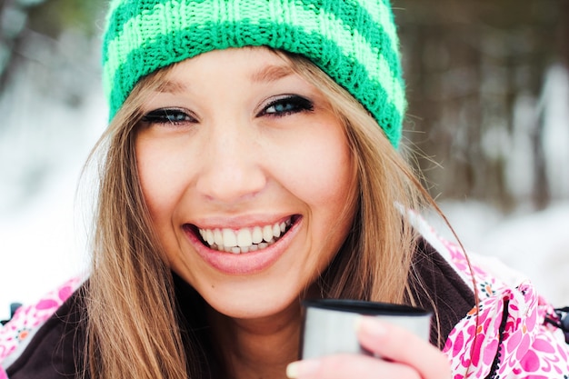 Jong mooi lachend meisje in een karmozijnrode jas en groene hoed die warme thee van een thermos in besneeuwde bergen drinken