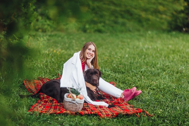 Jong mooi meisje brengt haar tijd door in het park met haar bruine hond zittend op het dekentapijt